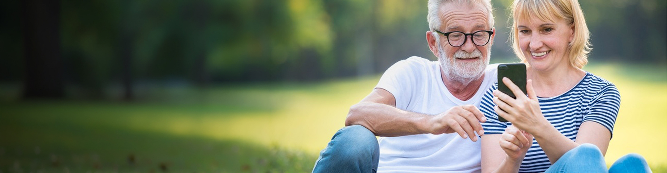 mature couple sitting in park on a sunny day looking at a mobile phone and smiling